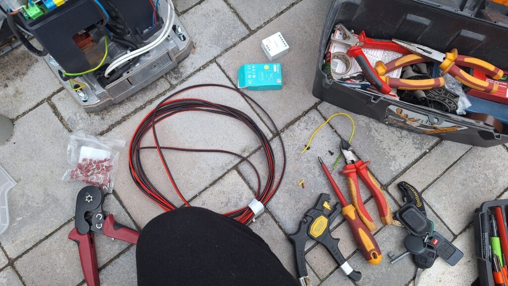 Villanyszerelői szerszámok és vezetékek elrendezve munkaterületen: piros és fekete kábelek, csavarhúzók, fogók, sorkapcsok, szerszámosláda és kapunyitó vezérlőmodul.